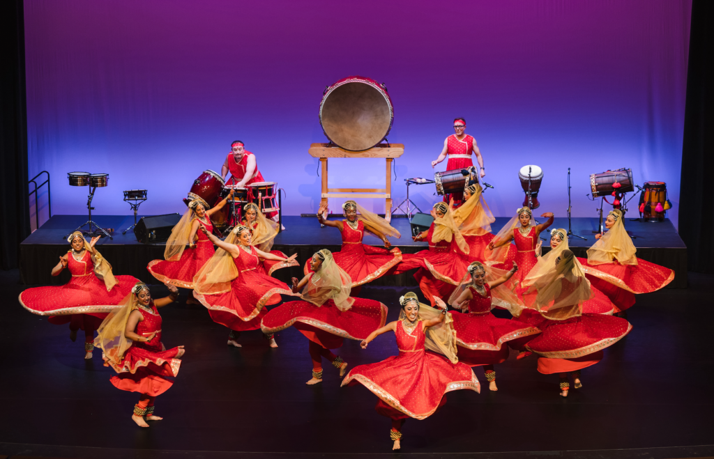 Mudra Dance Studio's performing company dancing, their beautiful costumes creating a wonderful circular tapestry as they spin togther. A taiko player and dholi are on a riser behind them.